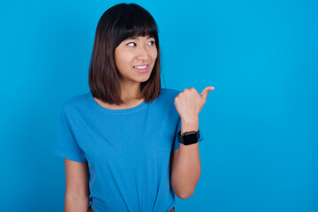 Fototapeta premium young beautiful asian girl wearing blue t-shirt against blue background points away and gives advice demonstrates advertisement