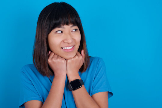 Young Beautiful Asian Girl Wearing Blue T-shirt Against Blue Background Holds Hands Under Chin, Glad To Hear Heartwarming Words From Stranger