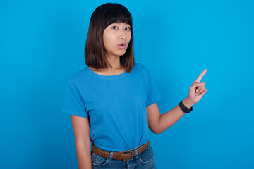 Emotive young beautiful asian girl wearing blue t-shirt against blue  keeps jaw dropped from shock demonstrates amazing promo points right on blank space demonstrates big shopping sale. Advertisement