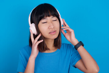Fototapeta premium young beautiful asian girl wearing blue t-shirt against blue background with headphones on her head, listens to music, enjoying favourite song with closed eyes, holding hands on headset.