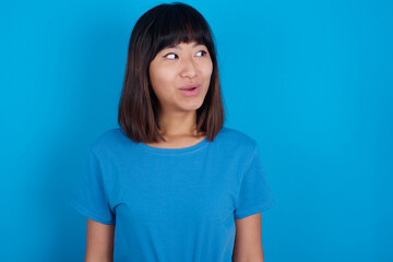 Shocked young beautiful asian girl wearing blue t-shirt against blue background look empty space with open mouth screaming: Oh My God! I can't believe this.