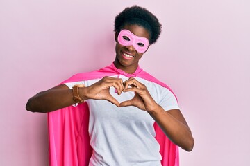 Fototapeta premium Young african american girl wearing superhero mask and cape costume smiling in love doing heart symbol shape with hands. romantic concept.