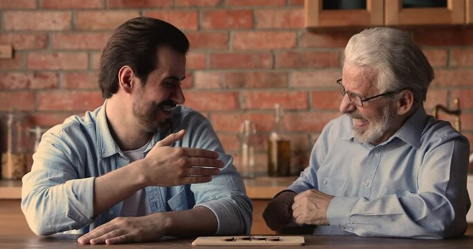 Cheerful elderly dad play checkers with adult grown up son sit at table in kitchen, young man makes final move win handshake to rival. Family have fun enjoy board game. Hobby, communication concept