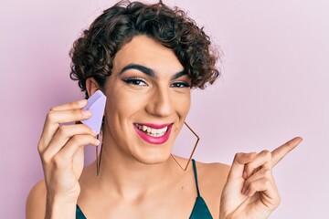 Young man wearing woman make up holding makeup sponge smiling happy pointing with hand and finger to the side