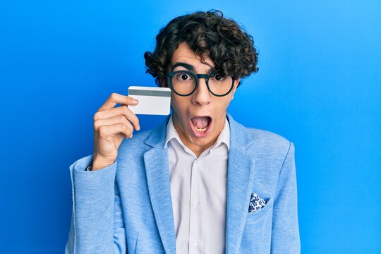 Hispanic young man holding credit card scared and amazed with open mouth for surprise, disbelief face