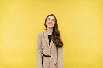 Fototapeta premium Portrait of a joyful woman with braces on her teeth emotionally smiling on a yellow background and looking at the camera, wearing a beige jacket