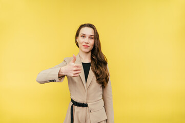 Positive female office worker in suit stands on yellow background, looks at camera with smile on face and shows thumbs up