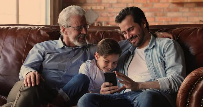 Three gen of men sit on couch at home laughing while watch funny online videos, little boy his young dad and older granddad spend time together using smartphone mobile apps. Virtual amusements concept