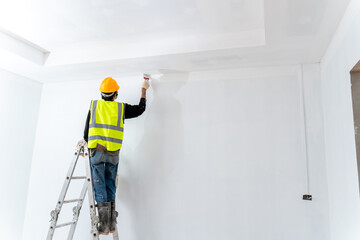 asian painter man painting the wall in an unfinished building, with paint brush and bucket, isolated on big empty space with ladder at the construction site