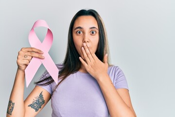 Young hispanic woman holding pink cancer ribbon covering mouth with hand, shocked and afraid for mistake. surprised expression