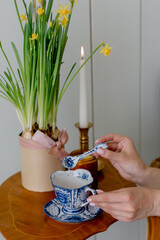Beautiful vintage table against the background of a green wooden wall of planks Yellow daffodils in a pot Long candle is burning Female hands are holding a porcelain spoon Cup is painted with a color