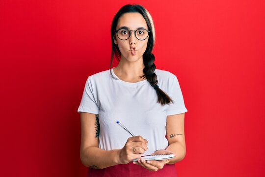 Young hispanic woman wearing waitress apron taking order making fish face with mouth and squinting eyes, crazy and comical.