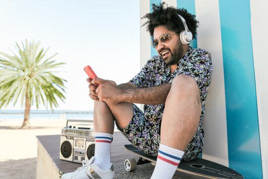 Afro Latin man having fun with mobile smartphone and listening music with headphones and vintage boombox stereo on tropical beach during vacation time
