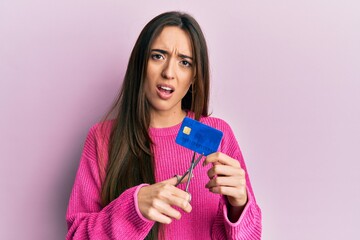 Young hispanic girl cutting credit card using scissors in shock face, looking skeptical and sarcastic, surprised with open mouth