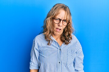 Middle age blonde woman wearing casual clothes and glasses in shock face, looking skeptical and...