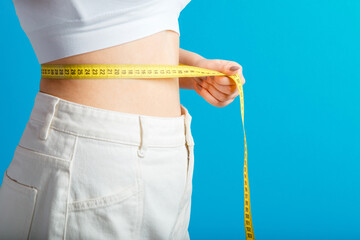 Slim woman measures her waist waistline with measuring tape. Healthy body shaping weight loss concept. Slim waist small belly in big white denim pants isolated over blue color background. Copy space