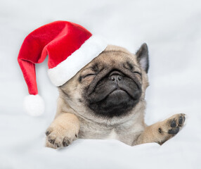 Funny Pug puppy wearing red santa hat sleeps under white blanket at home. Top down view