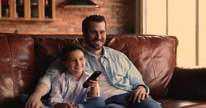 Little boy hold remote control switch channels relax watch online television together with young dad, family spend weekend at home talking discuss program while sit on sofa. TV lovers, leisure concept