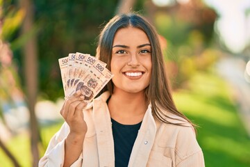 Young hispanic girl smiling happy holding mexican 500 pesos banknotes at the park. - obrazy, fototapety, plakaty