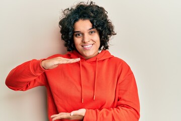 Young hispanic woman with curly hair wearing casual sweatshirt gesturing with hands showing big and large size sign, measure symbol. smiling looking at the camera. measuring concept.