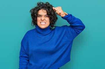 Fototapeta na wymiar Young hispanic woman with curly hair wearing turtleneck sweater angry and mad raising fist frustrated and furious while shouting with anger. rage and aggressive concept.
