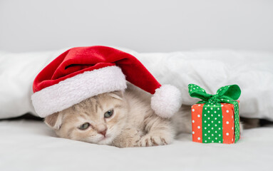 Sad kitten wearing red santa's hat lying with gift box under a white blanket on a bed