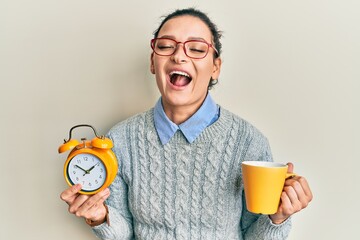 Young caucasian woman holding alarm clock drinking coffee smiling and laughing hard out loud because funny crazy joke.
