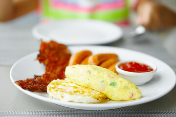 fried egg omelet grilled bacon and sausage with ketchup or tomato sauce on white dish for breakfast or lunch with hand of child or kid holding spoon to morning food eating at home or restaurant