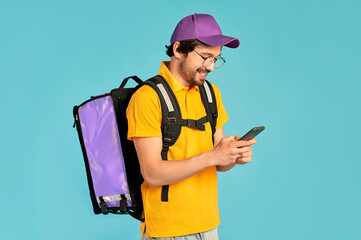 Portrait of a young courier, delivery man in uniform with a thermo backpack and a smartphone isolated on a blue background. Fast home delivery. Online order.