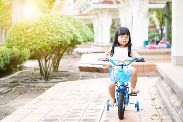 Fototapeta na wymiar Asian child preschool or kid girl wearing white shirt and cycling on blue bike or riding bicycle and happy on public park with green garden and tree to enjoy sport exercise with fun on summer holiday