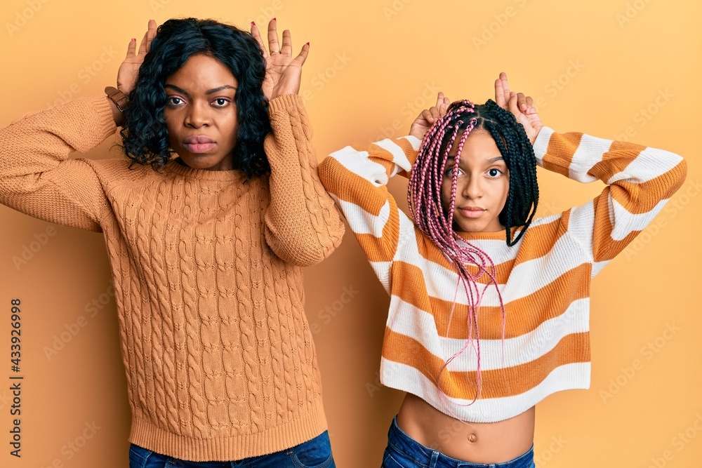 Poster Beautiful african american mother and daughter wearing wool winter sweater doing bunny ears gesture with hands palms looking cynical and skeptical. easter rabbit concept.