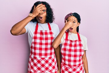 Beautiful african american mother and daughter wearing baker uniform peeking in shock covering face and eyes with hand, looking through fingers afraid