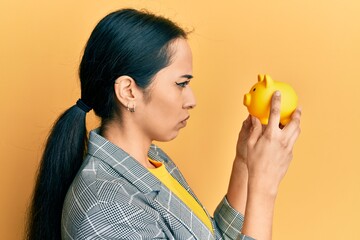Young hispanic girl looking to piggy bank skeptic and nervous, frowning upset because of problem....