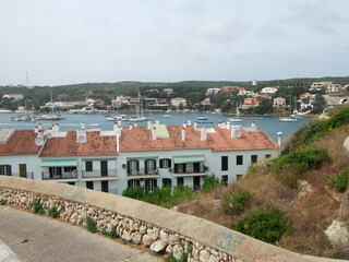 Fototapeta na wymiar View from a hill above residential houses to the small harbor of Menorca with many sailboats and fishing boats lying in the water of the ocean. In the background you can see beach houses on a hill