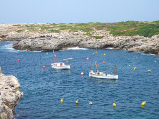 Small white wooden fishing boats are moored on the rocky coast of Menorca in front of some colorful buoys. Some rocky edges washed out by the sea can be seen