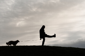 Silhouette of a man on top of a hill at sunset doing a silly walk. His dog, on leash, is behind him.   - obrazy, fototapety, plakaty