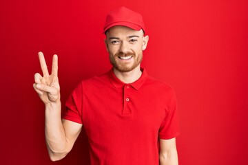 Young redhead man wearing delivery uniform and cap smiling with happy face winking at the camera doing victory sign. number two.