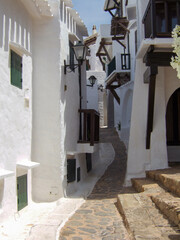 Typical Spanish construction of a narrow alley between white beach houses built according to Cycladic architecture in Menorca. To the right and left are some balconies with wooden railings.