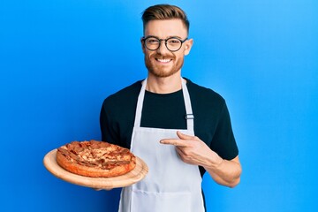 Young redhead man wearing waiter apron holding pizza smiling happy pointing with hand and finger