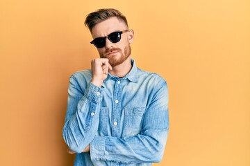 Fototapeta premium Young redhead man wearing stylish sunglasses serious face thinking about question with hand on chin, thoughtful about confusing idea
