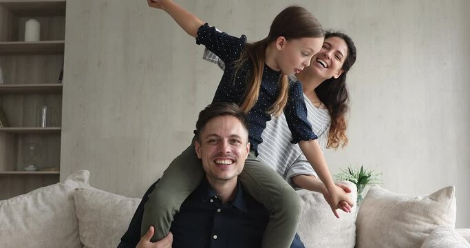 Latin married couple and their daughter having fun at home, girl sit on dad shoulders holding hands loving mother spreading arms imagining flying laughing enjoy playtime. Happy family portrait concept