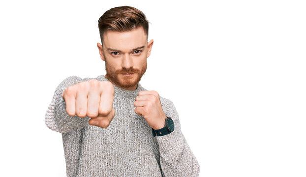 Young redhead man wearing casual winter sweater punching fist to fight, aggressive and angry attack, threat and violence