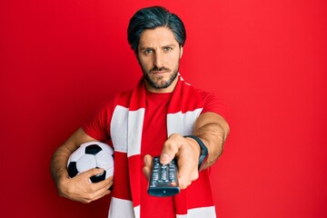 Young hispanic man football hooligan holding ball using tv control skeptic and nervous, frowning upset because of problem. negative person.