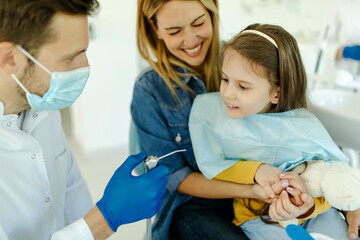 Obraz premium Little cute smiling girl is sitting in dental chair with mother in clinic, office.