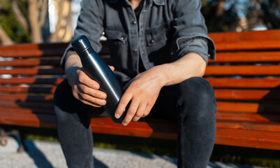 Close-up of male hands, holding steel reusable thermo water bottle of black. Man sitting on bench...