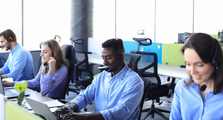 Smiling african male call-center operator with headphones sitting at modern office with collegues on the backgroung, consulting online.