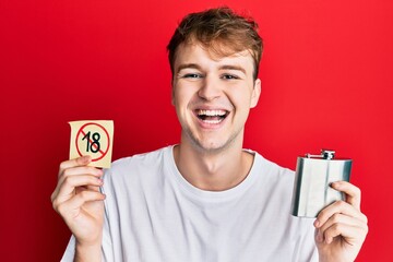 Young caucasian man holding whiskey flask and under 18 forbidden symbol smiling and laughing hard...