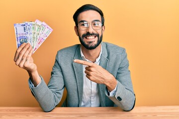 Young hispanic man holding indian rupee banknotes sitting on the table smiling happy pointing with hand and finger