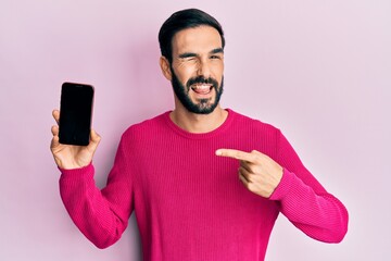 Young hispanic man holding smartphone showing blank screen winking looking at the camera with sexy...