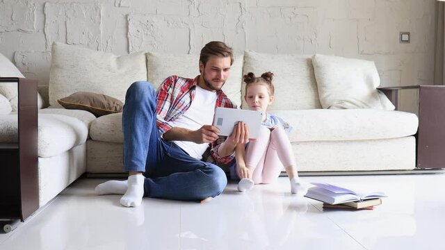Smiling father and daughter sitting on floor in living room with digital tablet, teaching lessons.